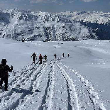 Mehrere Wanderer mit Stöcken auf verschneitem Bergpfad vor Alpenpanorama.