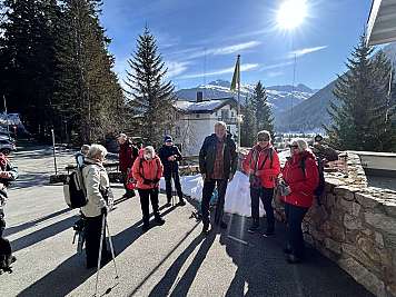Several hikers stand on a sunny mountain road with snow, trees, and peaks behind them.