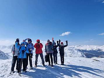 Gruppe von Skifahrern steht auf einem schneebedeckten Berggipfel und winkt in die Kamera.