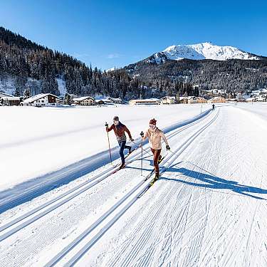 Zwei Langläufer auf präparierter Spur in einer verschneiten Berglandschaft.