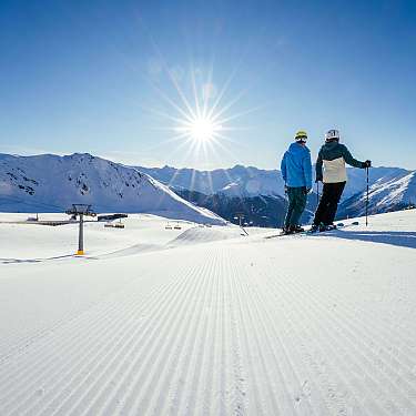 Zwei Skifahrer stehen auf einer präparierten Schneepiste mit Bergpanorama und Sonnenlicht.