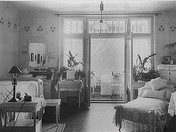A bright bedroom with several beds, mirrors, potted plants, and lace-covered furniture arranged around a large window