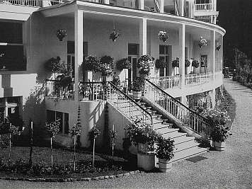 Curved white house exterior with stairs, balcony railings, and potted flowers beside a gravel path in black and white.
