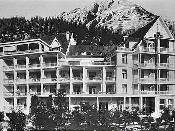Large alpine hotel with balconies in front of a snowy mountain and pine forest, black and white view of an expansive facade.