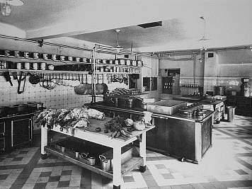 Black-and-white view of a large stainless-steel kitchen with hanging cookware and prep tables,