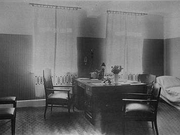 Dim office room with desk, chairs, bed, and striped wall panels in grayscale; no people visible.
