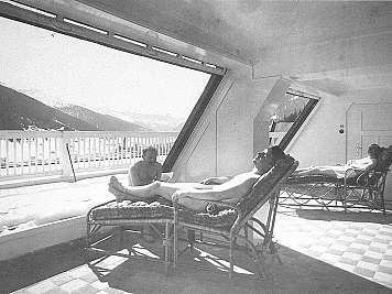 People reclining on lounge chairs in a bright terrace room overlooking snowy mountains and a balcony railing.,