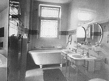 Black-and-white bathroom with bathtub, two pedestal sinks, round mirrors, and tiled walls with a window above the tub.,