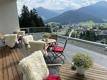 Outdoor terrace with wooden tables, fluffy chairs, and mountain village views