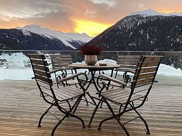 Wooden table and chairs on a snowy balcony overlooking mountains at sunset.