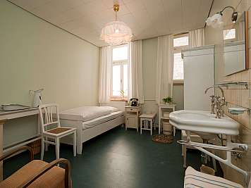 A tidy room with a bed, sink, chair, and window curtains.