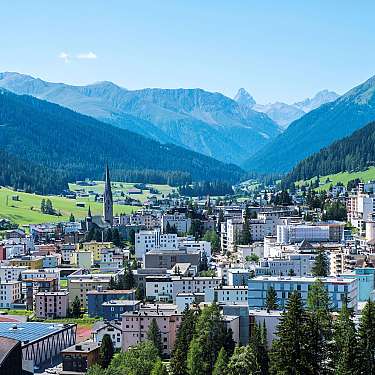 Blick auf eine Alpenstadt mit Kirchturm, Wohnhäusern und Bergen im Hintergrund.
