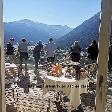 People stand on a rooftop terrace overlooking mountains, with breakfast drinks and pastries on a table.