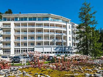 White multi-storey hotel building with balconies, surrounded by trees and parked cars