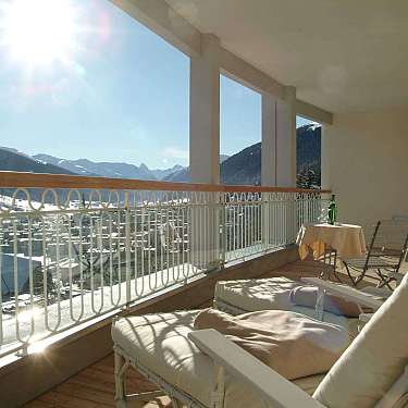 Balkon mit Sonnenliegen und Tisch, Blick auf verschneite Berge und Tal bei sonnigem Wetter