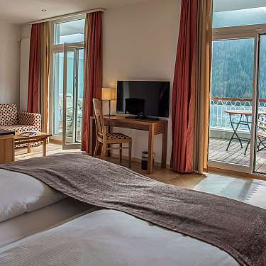 A hotel bedroom with balcony doors, TV, desk, and mountain view.