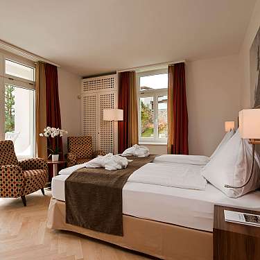 Bright hotel room with bed, armchairs, and mountain view through large windows.