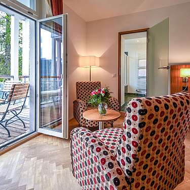 Helles Hotelzimmer mit Balkon, zwei gemusterten Sesseln, Bett und geöffneten Glastüren.