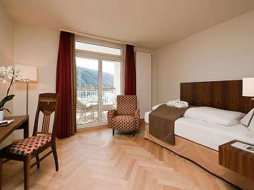 Modern hotel room with bed, balcony doors, patterned chairs, and mountain view through the window.