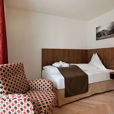 A modern hotel room with a bed, patterned armchair, wood headboard, and window drapes.