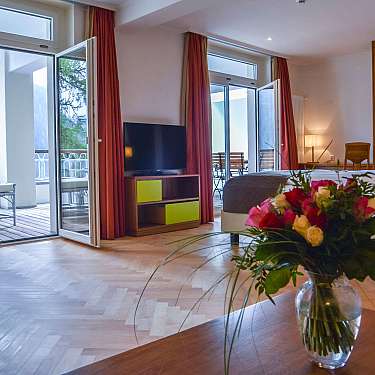Spacious hotel room with open balcony doors, bed, television, and flower bouquet on a table.