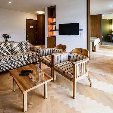 Spacious hotel suite with patterned sofa, wooden chairs, coffee table, and wall-mounted TV.
